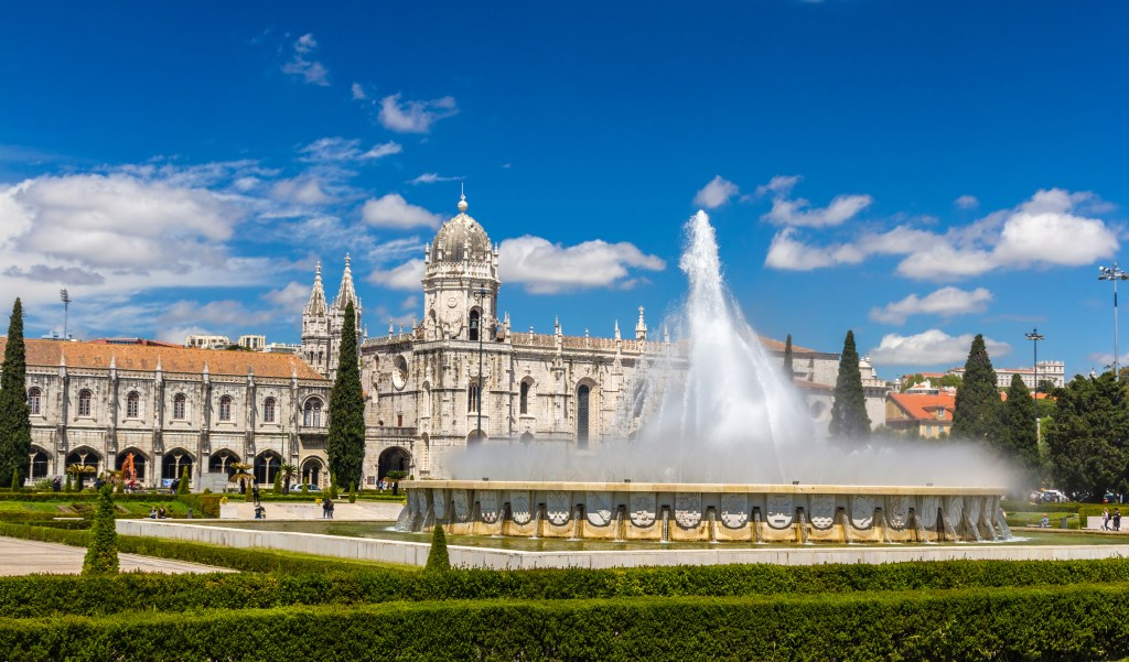 Manicured gardens and a fountain shooting water up into the air in front of an ornate building with a domed tower.